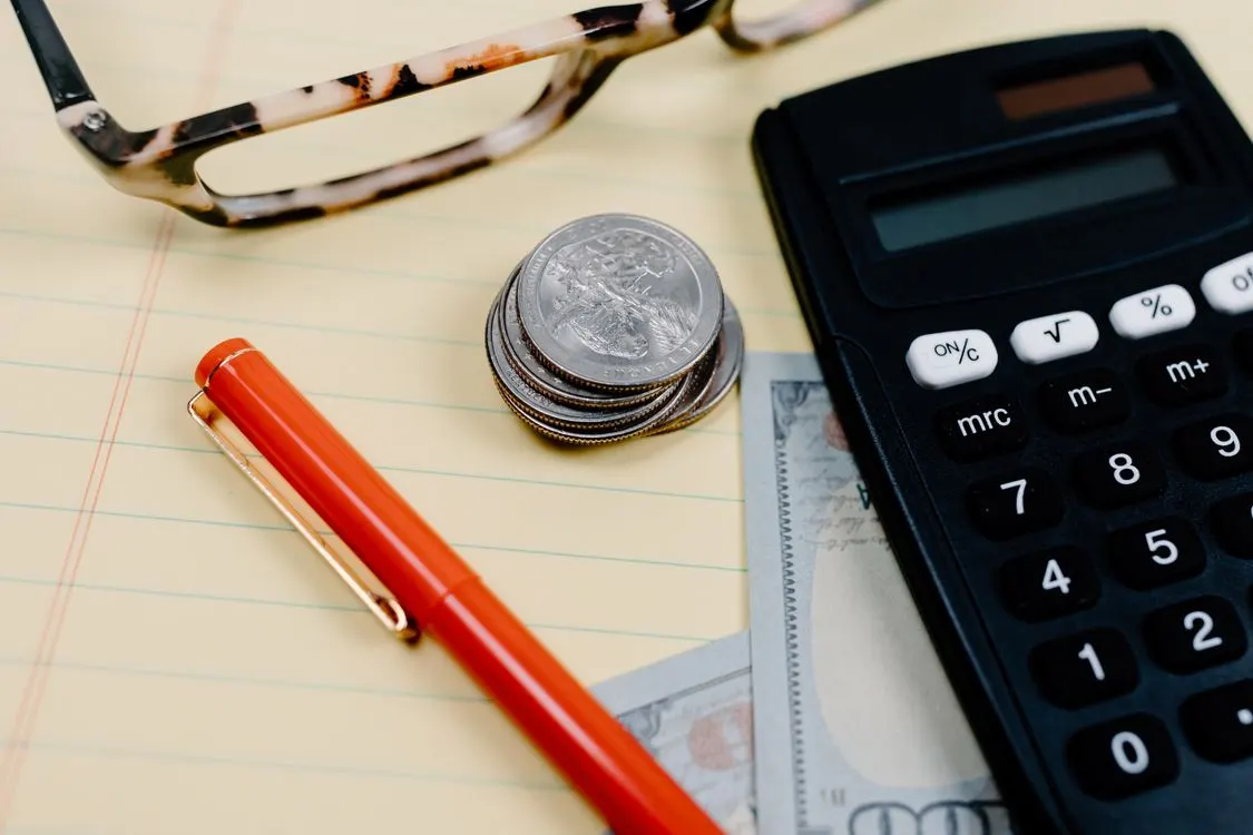 A calculator, coins and red pen beside an open notebook